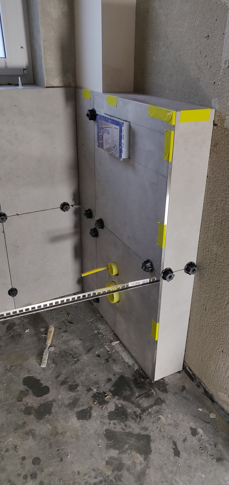 Badezimmer-Baustelle: Fliesenarbeiten, gelbe Montagescheiben, Keile und Metall-Schiene installiert.