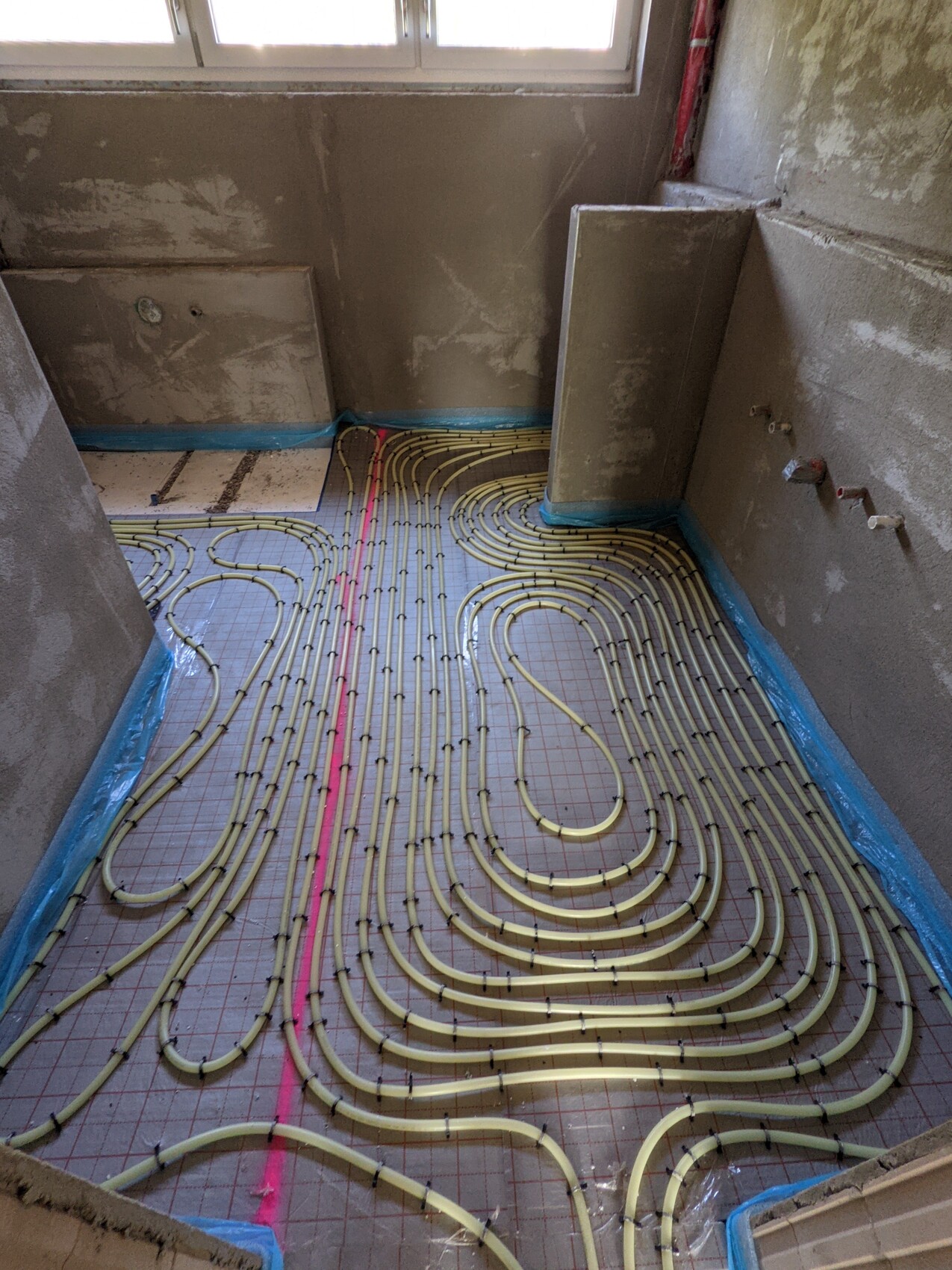 Unrenoviertes Badezimmer im Bau: Gelbe Fußbodenheizungsrohre in Schleifen auf einer Dämmplatte verlegt.