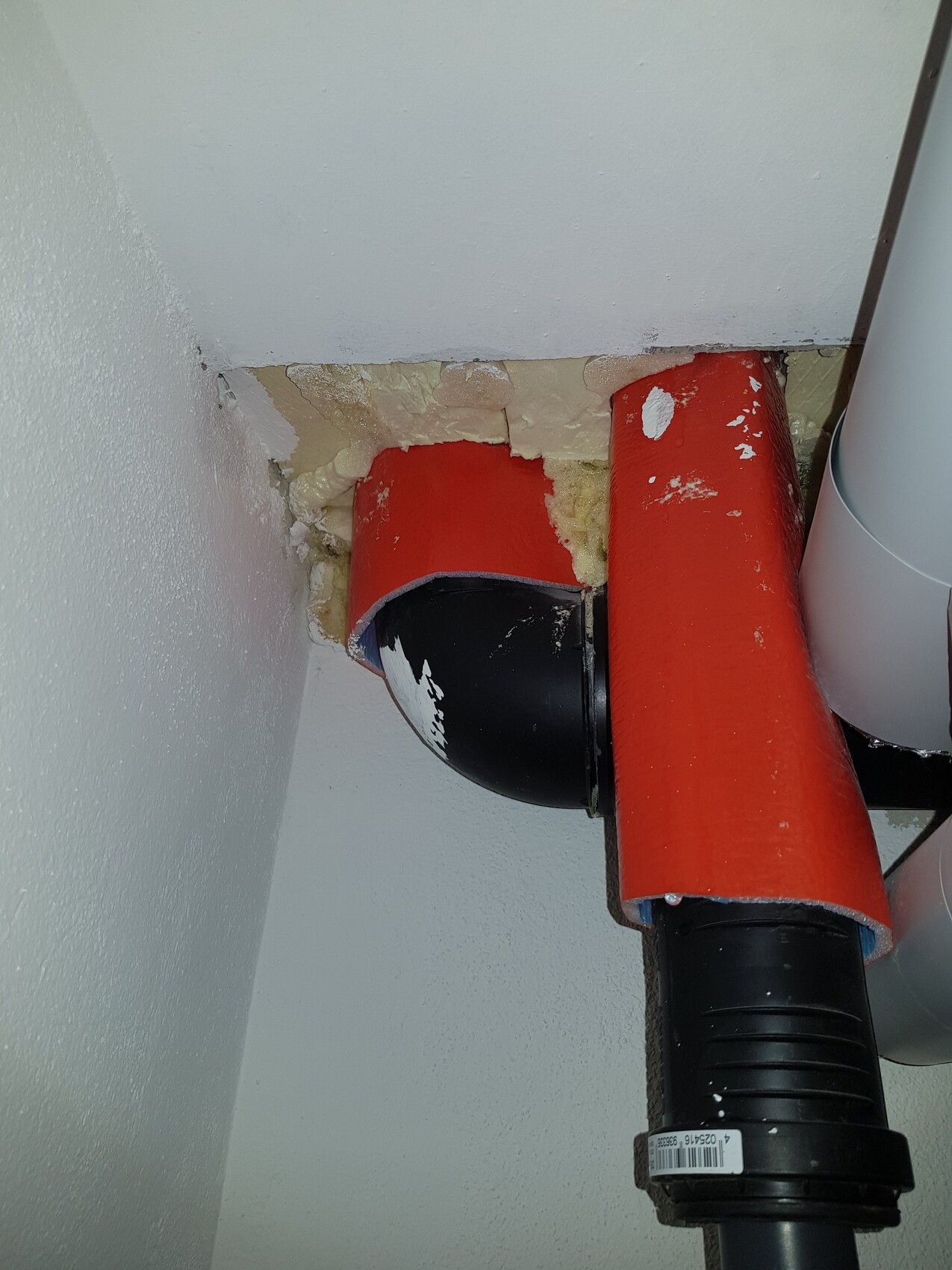 Rotes PVC-Rohr mit schwarzem Bogen führt durch Loch in Decke/Wand; umlaufender gelber Dämmschaum.