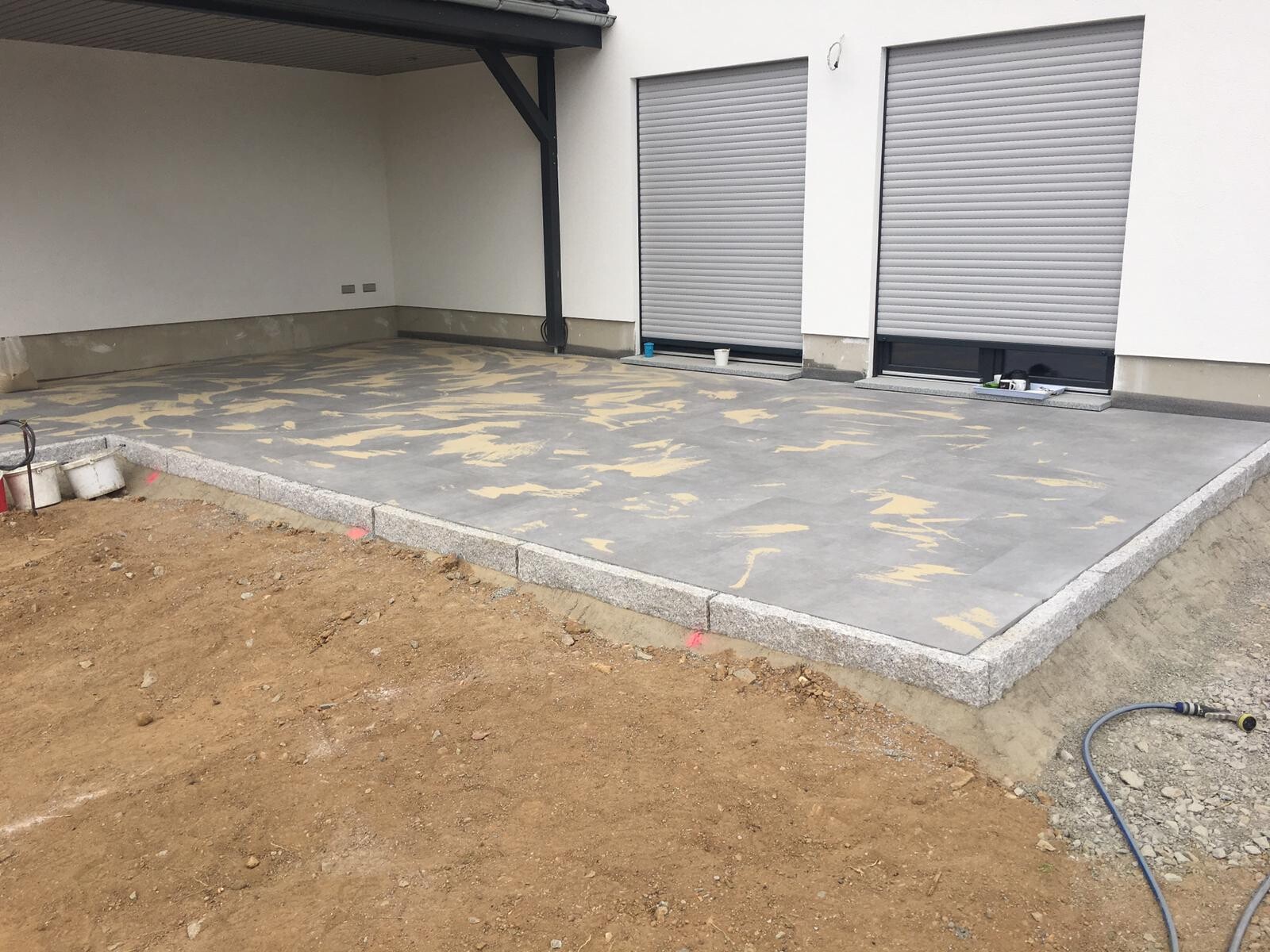 Neue Beton-Terrasse mit Granitrand vor dem Haus; zwei geschlossene Rollläden.