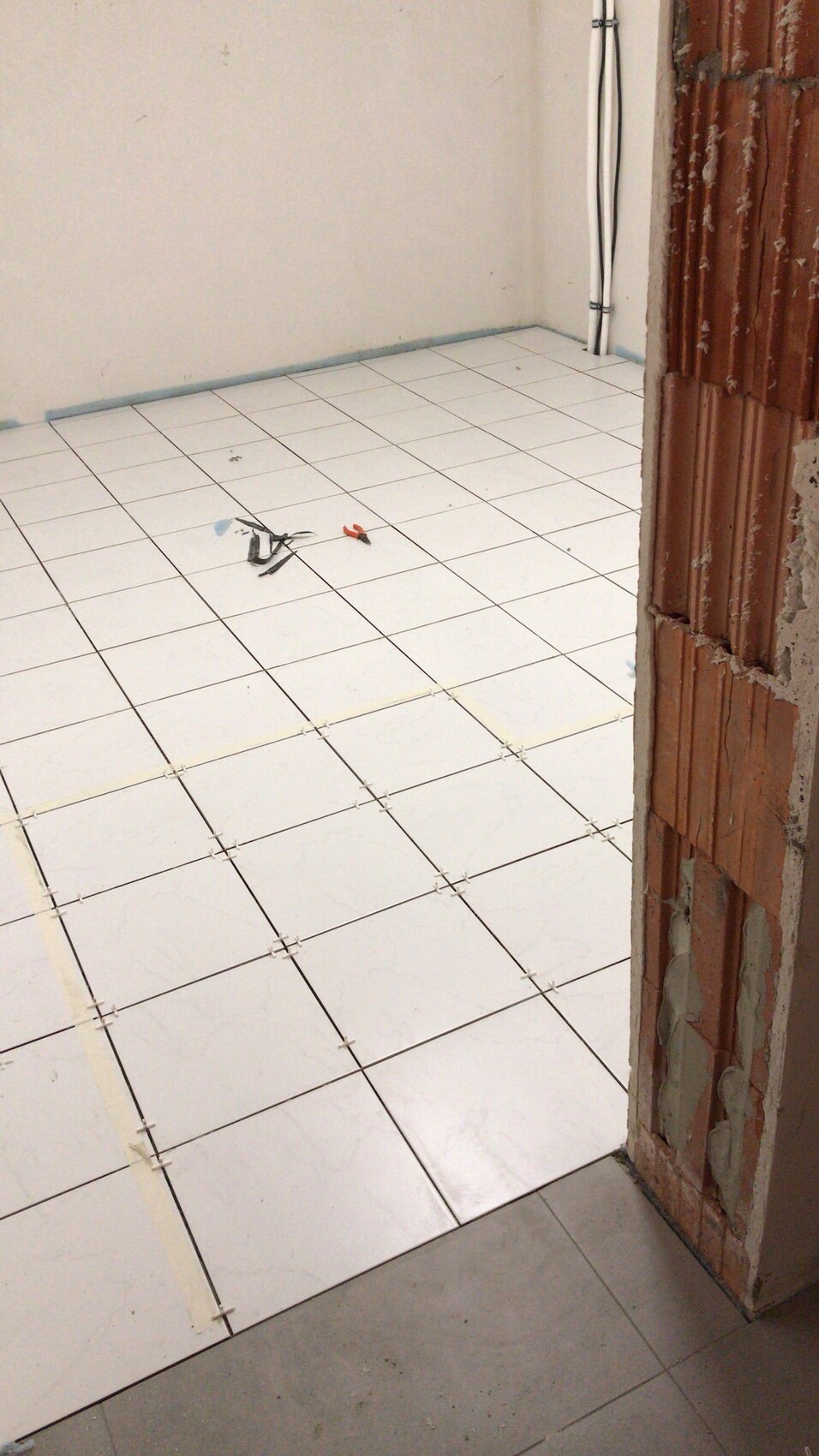 Bauzimmer mit weißem Fliesenboden, Fliesenclips sichtbar, offene Ziegelwand rechts.