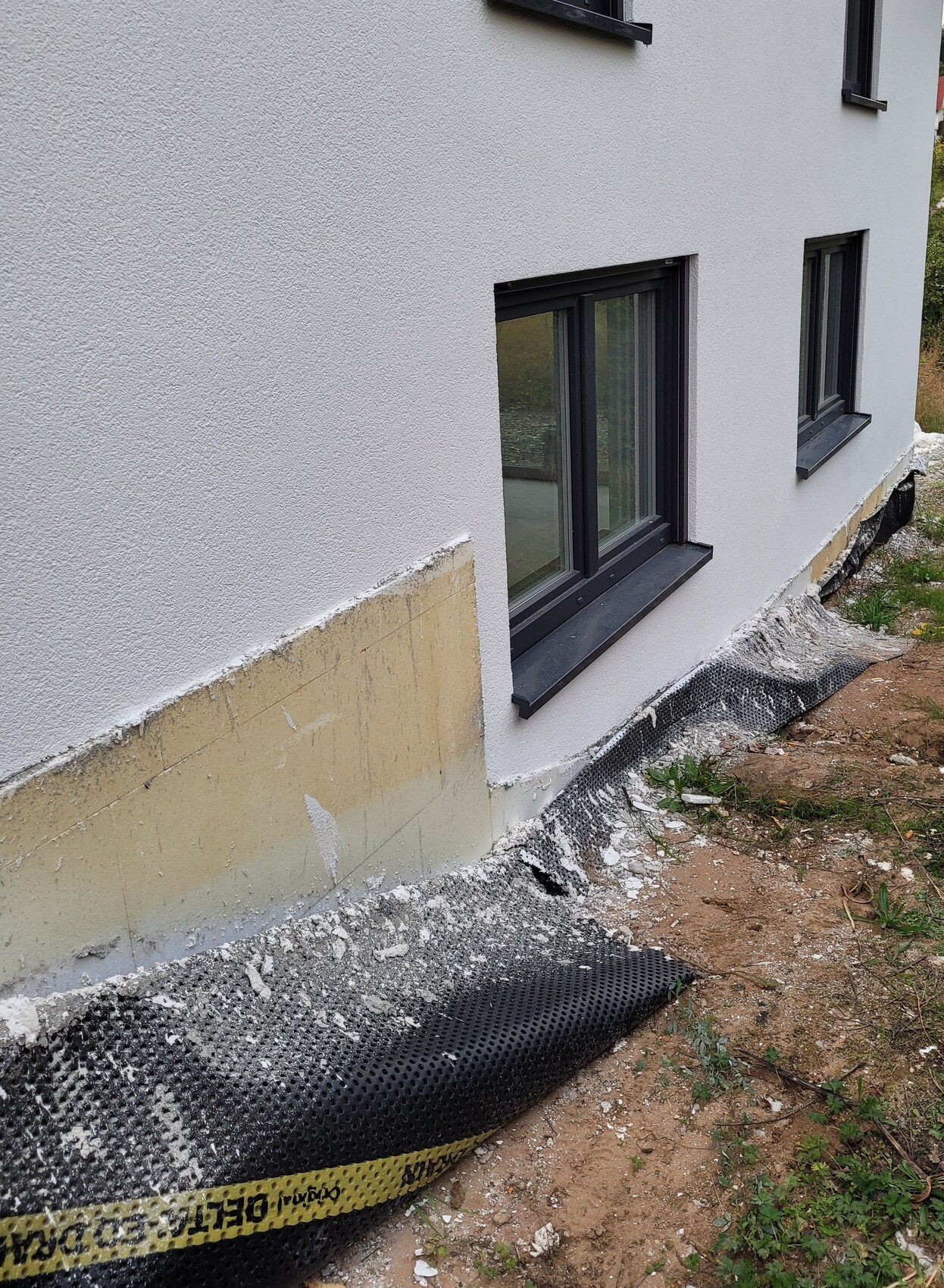 Außenwand eines Hauses mit grauem Putz, dunklen Fensterrahmen, Absperrband und Fundamentarbeiten.