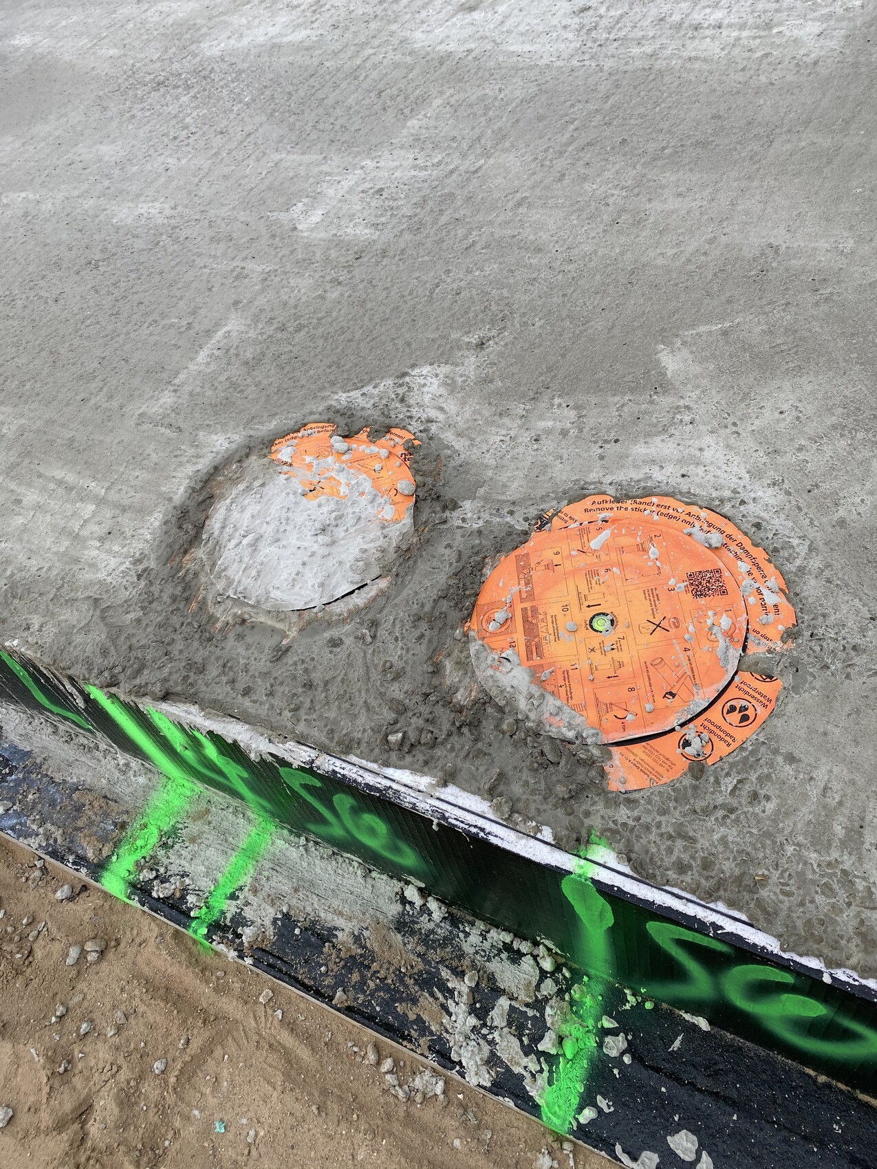 Zwei runde Deckel in frischem Beton einer Baustelle, orange und grau, neben grüner Bemalung.