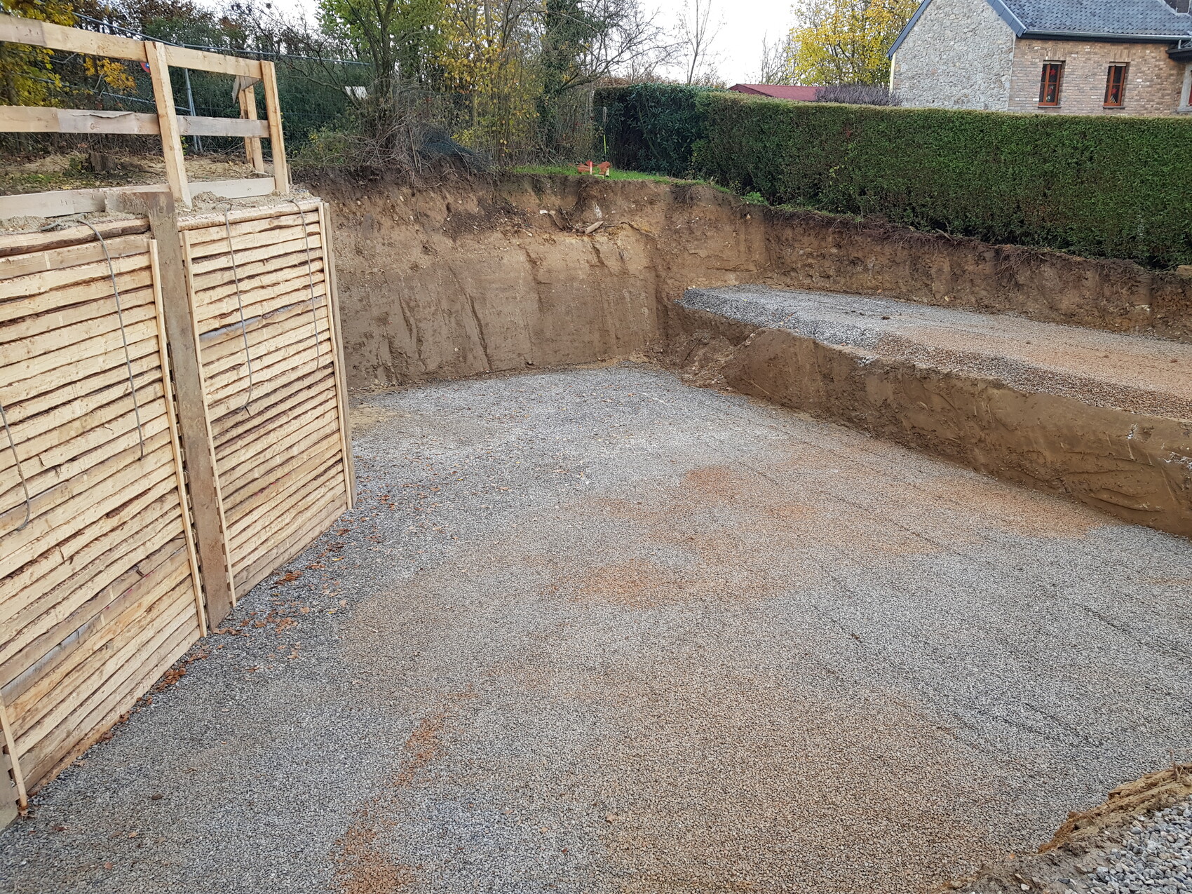 Baustelle mit Graben, Holzpalisade, Kiesboden, Hecke und Haus im Hintergrund
