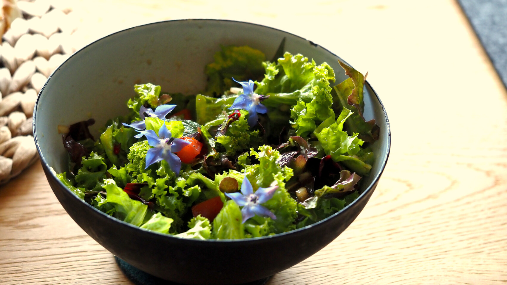 Schüssel mit grünem Salat, roten Tomaten und blauen Blüten auf Holztisch.