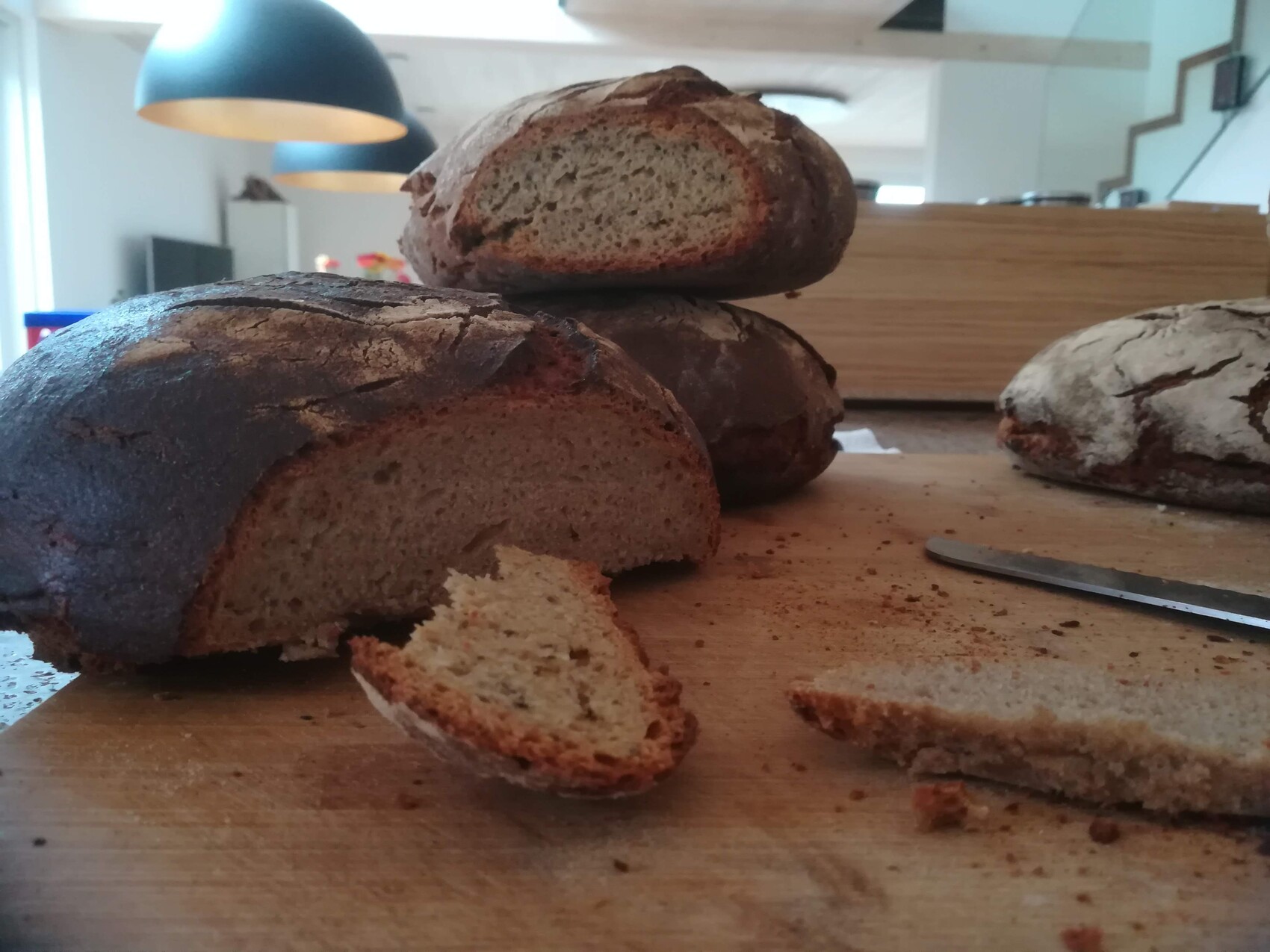Dunkles Brot auf Holzbrett; Scheibe abgebrochen, Messer daneben, Küchenhintergrund.