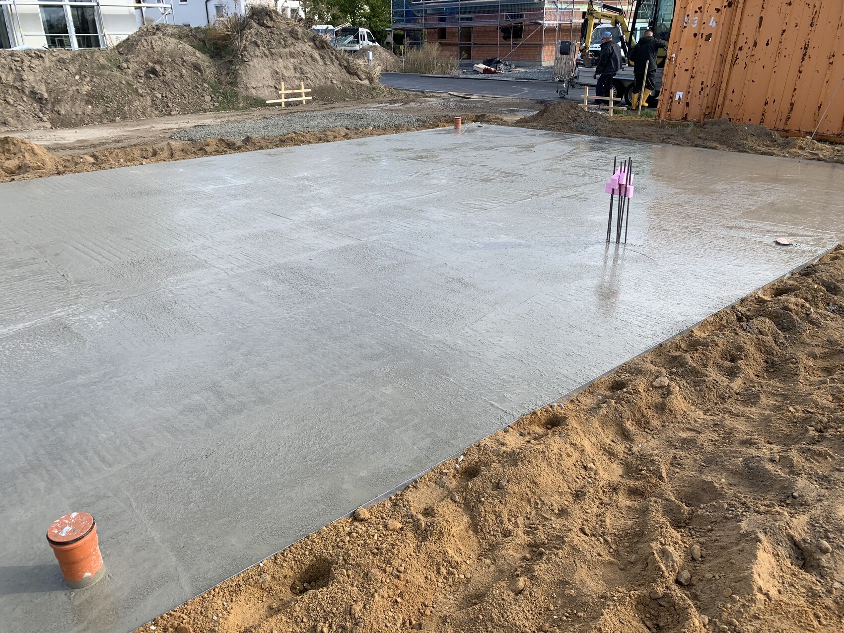 Frisch gegossene Betonbodenplatte auf Baustelle, Bewehrung ragt aus dem Beton, Erde umliegt.