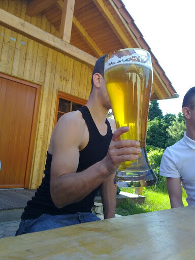 Mann in schwarzem Tanktop hält großes Bierglas vor das Gesicht im Freien neben einer Holzhütte.
