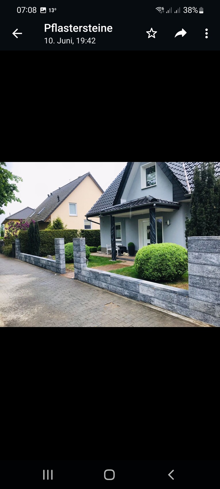 Vorderansicht eines grauen Einfamilienhauses mit Veranda, Säulen, Steinmauer und Hecken.