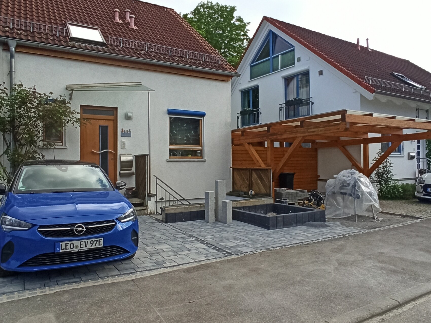 Blaues Auto vor weißem Haus; rechts eine Holzterrasse/Pergola und Baumaterialien.