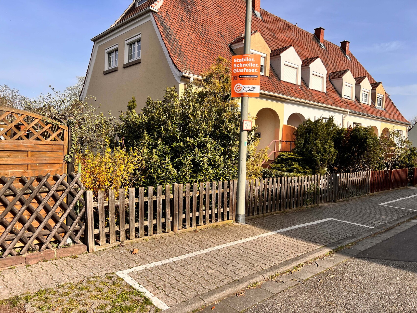 Straßenzug mit Häusern, rote Ziegeldächer, Holzzaun und orange Werbetafel am Pfosten