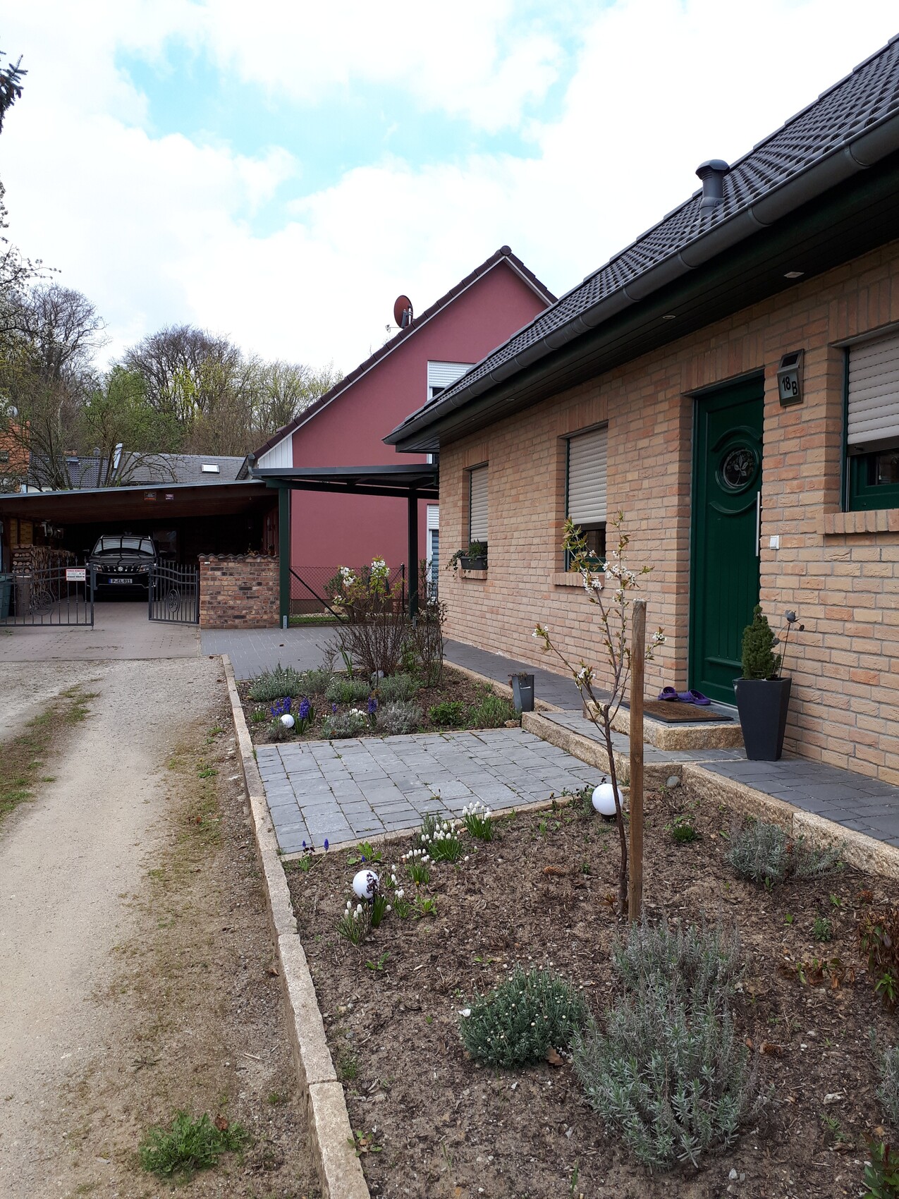 Vorderhaus aus Ziegeln mit grüner Tür, kleiner Garten, gepflasterter Weg, Carport im Hintergrund.