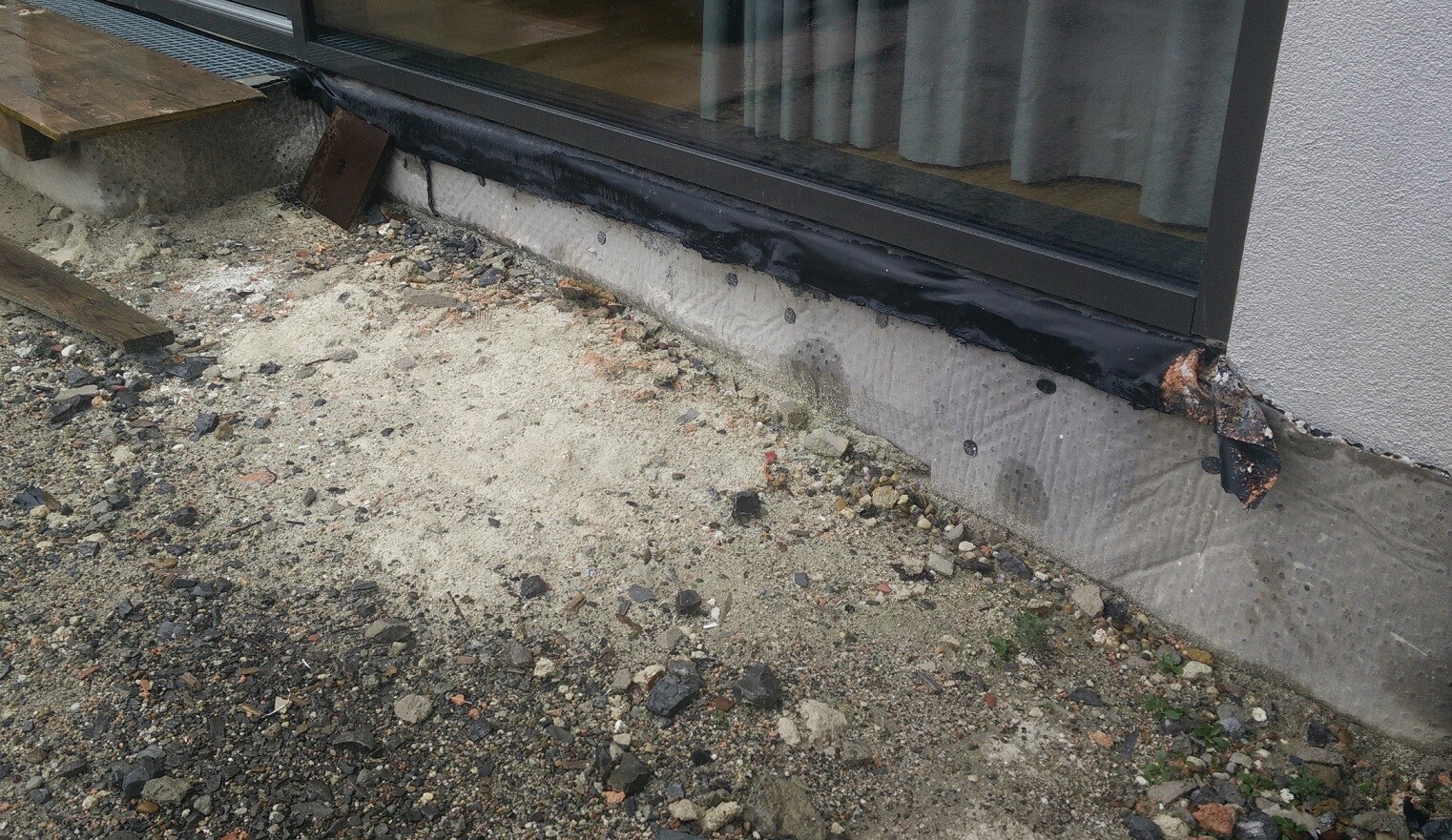 Außenansicht: Sockelabdichtung beschädigt, Kies- und Sandboden vor Glasfront.