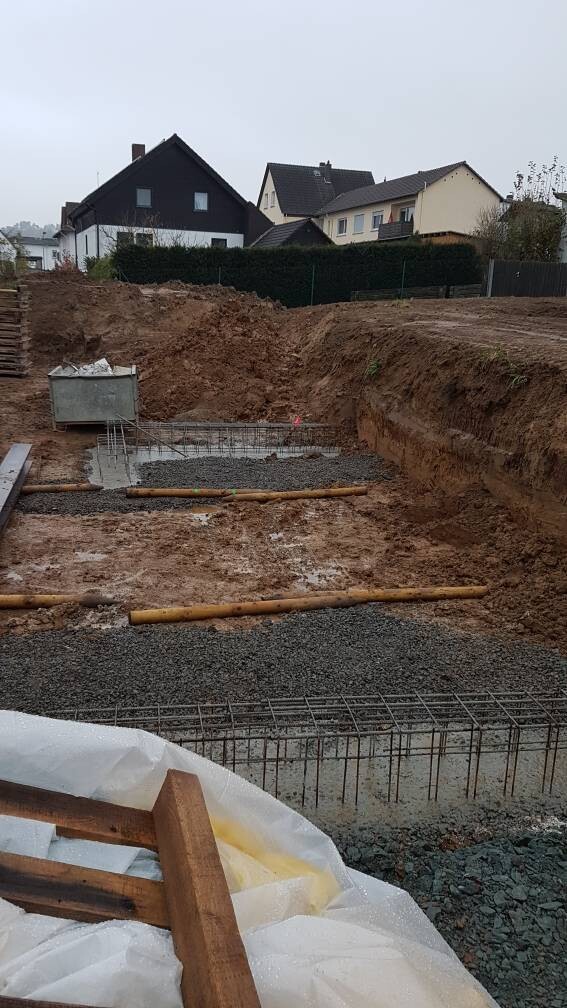 Baustelle mit ausgegrabener Baugrube, Bewehrungsgitter, Kies und Holzbohlen, Häuser im Hintergrund