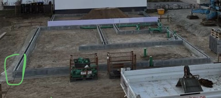 Baugrube mit Fundamentrahmen und Gräben auf einer Baustelle, Maschinen und Paletten im Hintergrund.