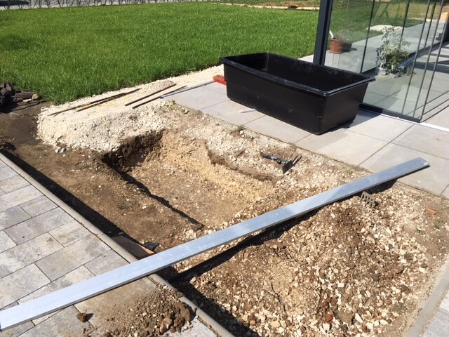 Baugrube an der Terrasse neben dem Haus, Kiesboden, Metallstange über Grube, schwarze Pflanzbox.