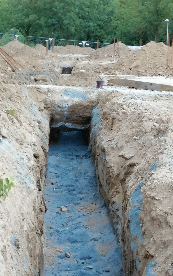 Graben auf Baustelle mit blauem Untergrund, Erdhaufen, Zaun und Wald im Hintergrund.