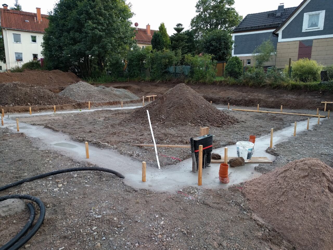 Baustellenfundament: Aushub, Kies- und Erdhaufen, Pfosten, Rohre, Eimer; Häuser im Hintergrund