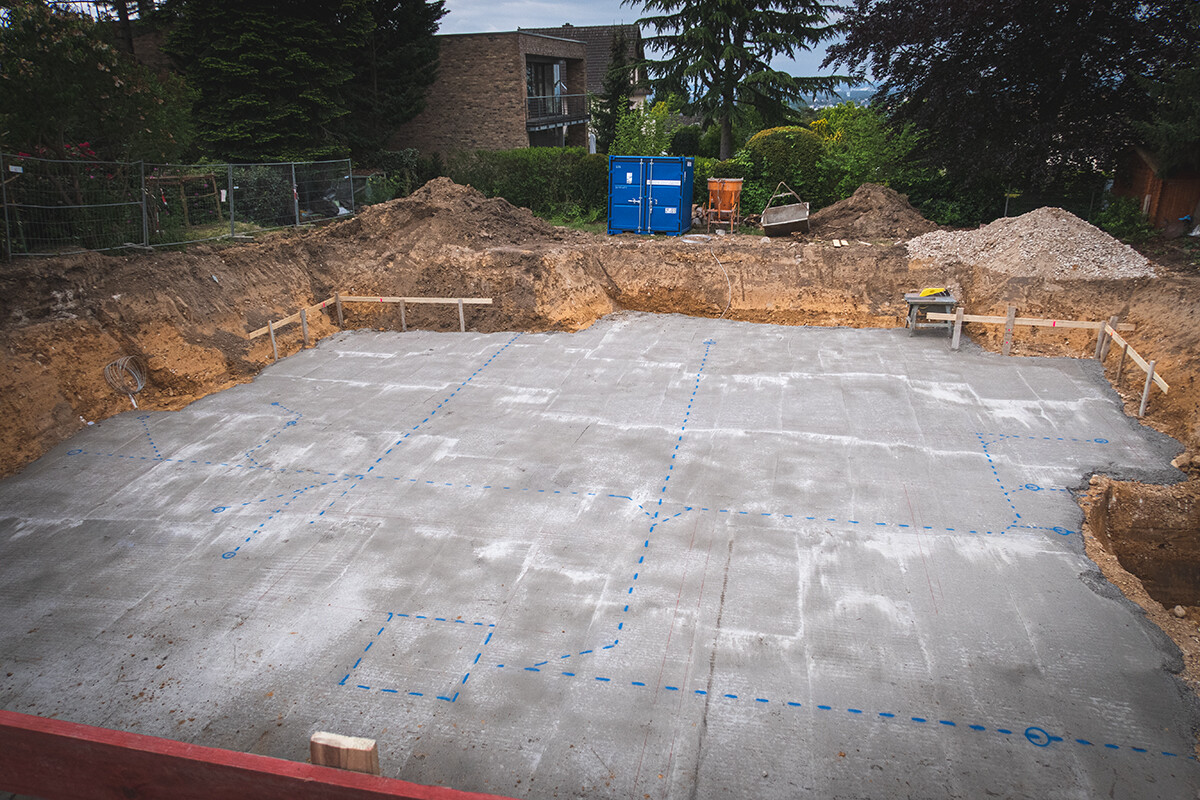 Aushub-Grundfläche: Betonplatte mit blauen Markierungen, Holzborde, Baucontainer im Hintergrund.
