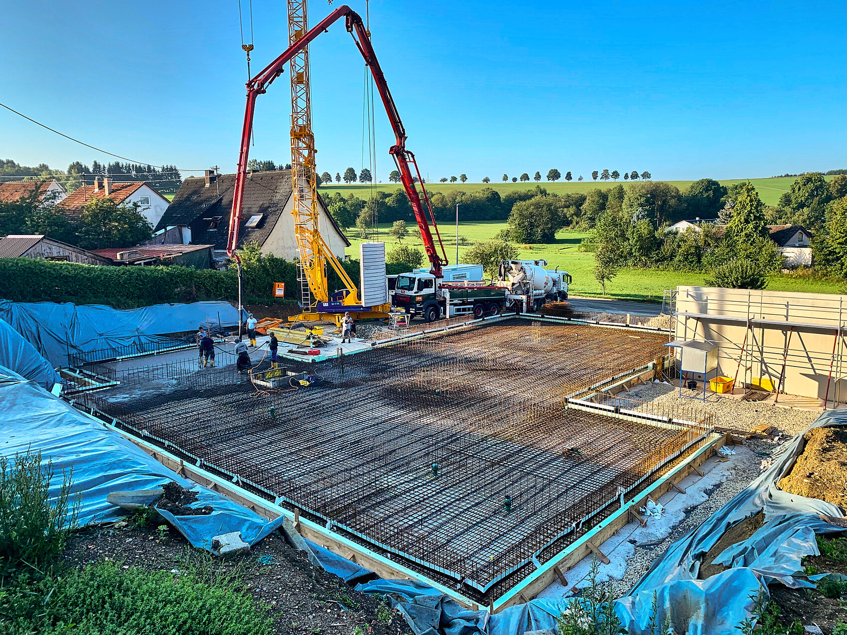 Große Baustelle mit Betonfundament, Stahlbewehrung, Kran und Arbeitern, ländliche Gegend.