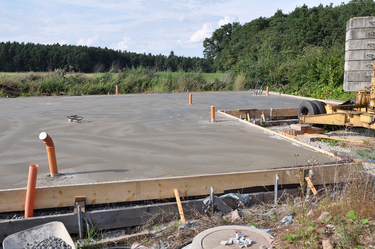 Große Betonplatte mit Holzformrahmen, orange Rohre ragen heraus, Baugerätschaften rechts.