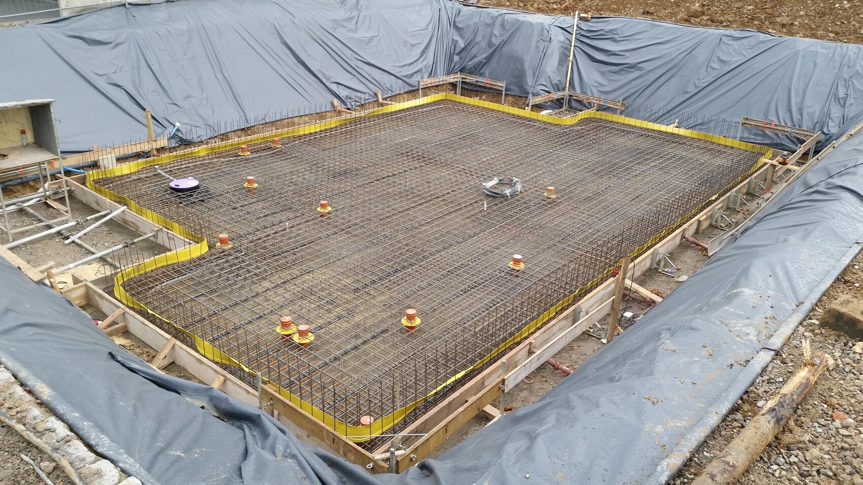 Fundament mit Bewehrungsstahl-Gitter in Baugrube, von Schutzplanen umgeben.