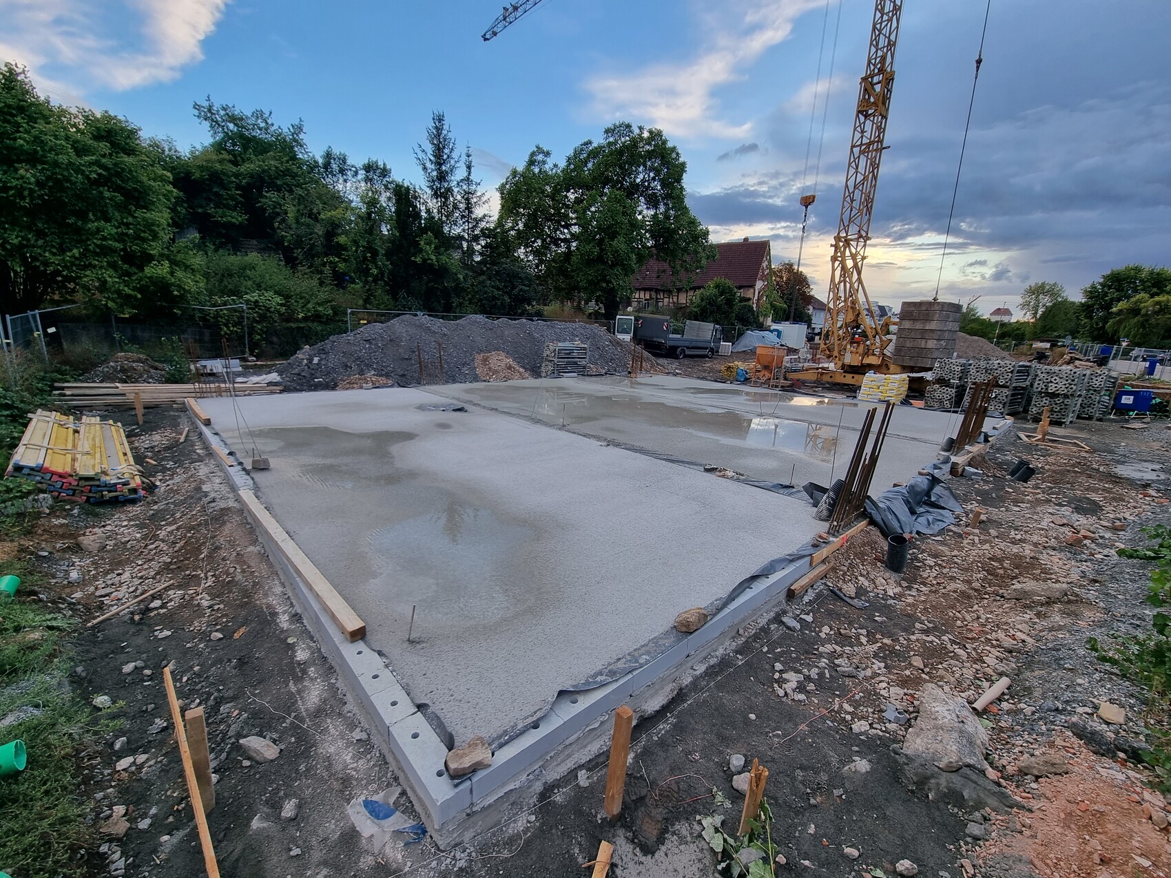 Baustelle mit Betonfundament, Kran, Holzstangen und Baustoffen im Hintergrund.