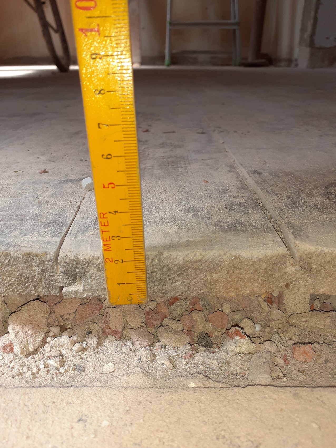 Gelbes Messband steht senkrecht neben einer Betonstufe, darunter Kies und Bruchreste.