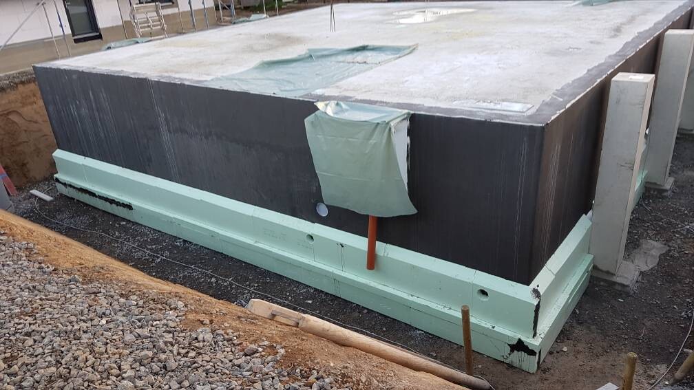 Großer schwarzer Betonblock auf grüner Dämmstoff-Platte; orangenes Rohr ragt hervor – Baustelle.