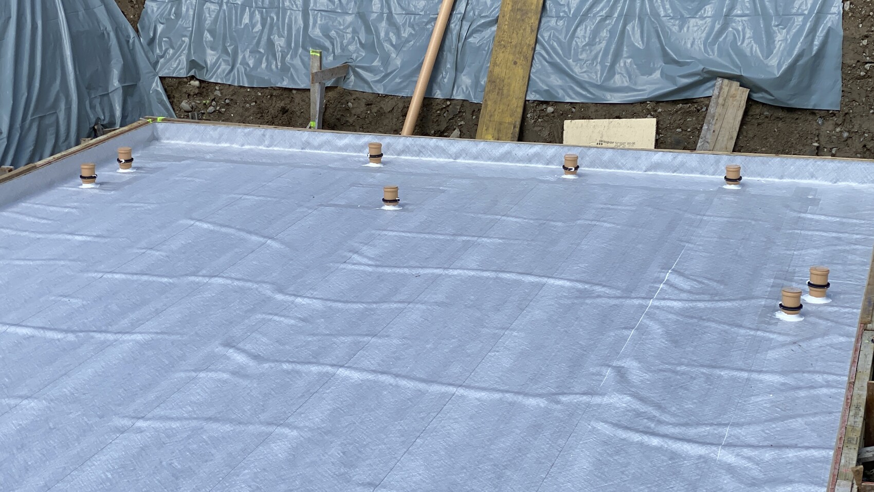 Fundamentplatte mit blauer Abdichtungsfolie, umlaufender Holzrahmen und Rohre mit Deckeln.