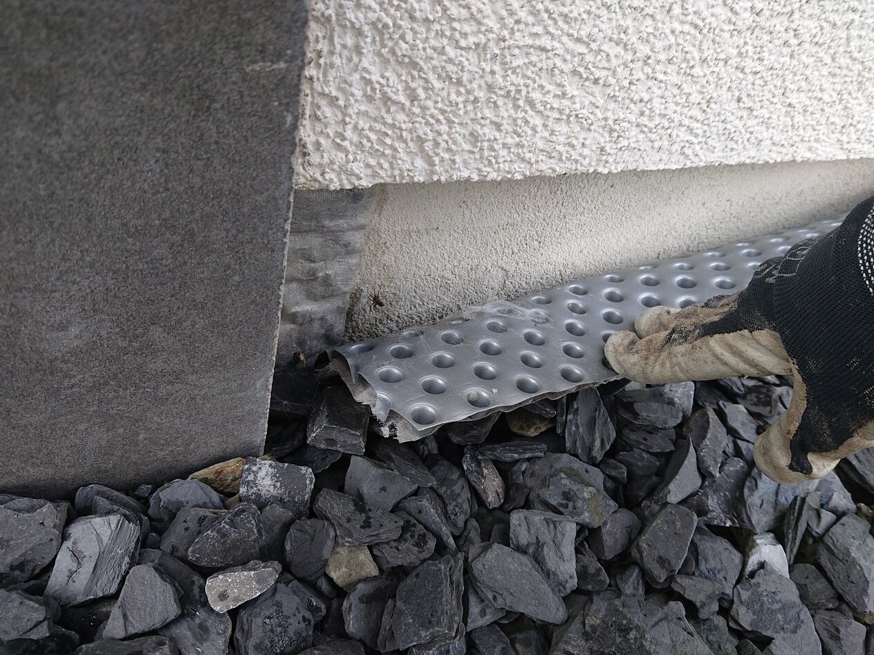 Hand mit Arbeitshandschuh legt perforierte Metallbahn gegen das Gebäudefundament über Kies.