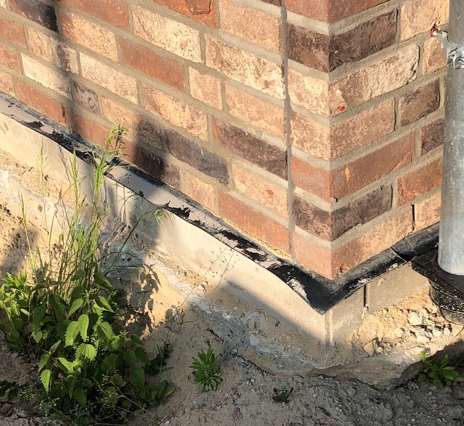 Eckbereich einer Ziegelwand mit Fundament, Abdichtung und Unkraut am Baugrund