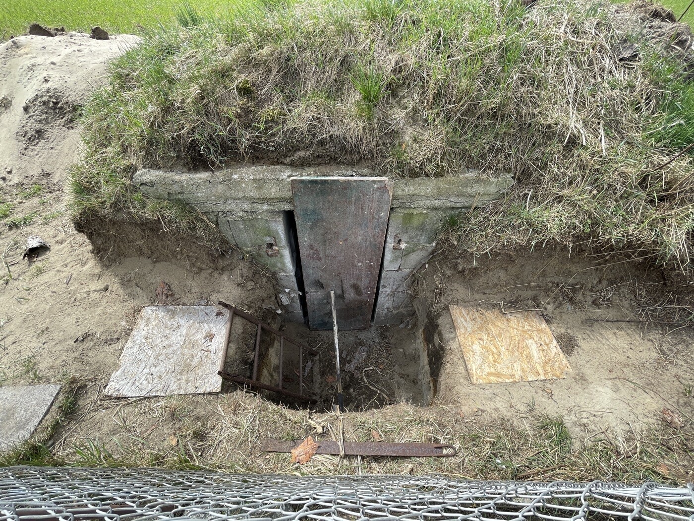 Grubenöffnung in Erdwall mit Betonrahmen, Metallklappe; rostige Leiter und Holzplatten davor.
