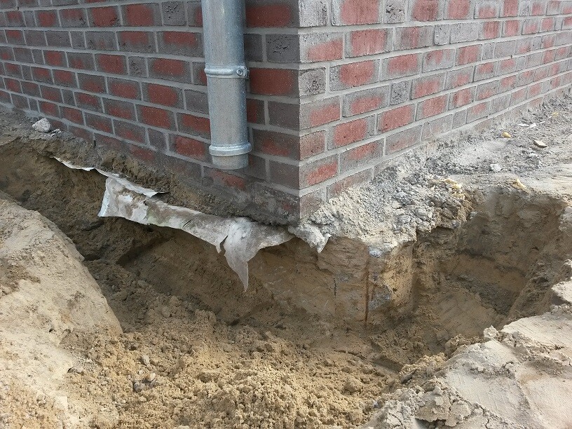 Fundamentbereich eines roten Backsteinhauses mit offenem Erdreich und Fallrohr an der Wand