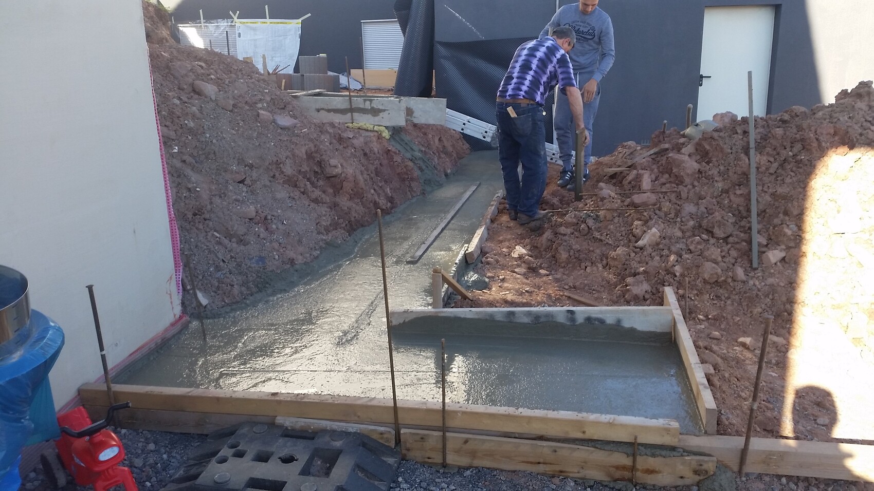 Zwei Bauarbeiter gießen frisches Betonfundament in eine Grabenform aus Holz, mit Bewehrungstäben.