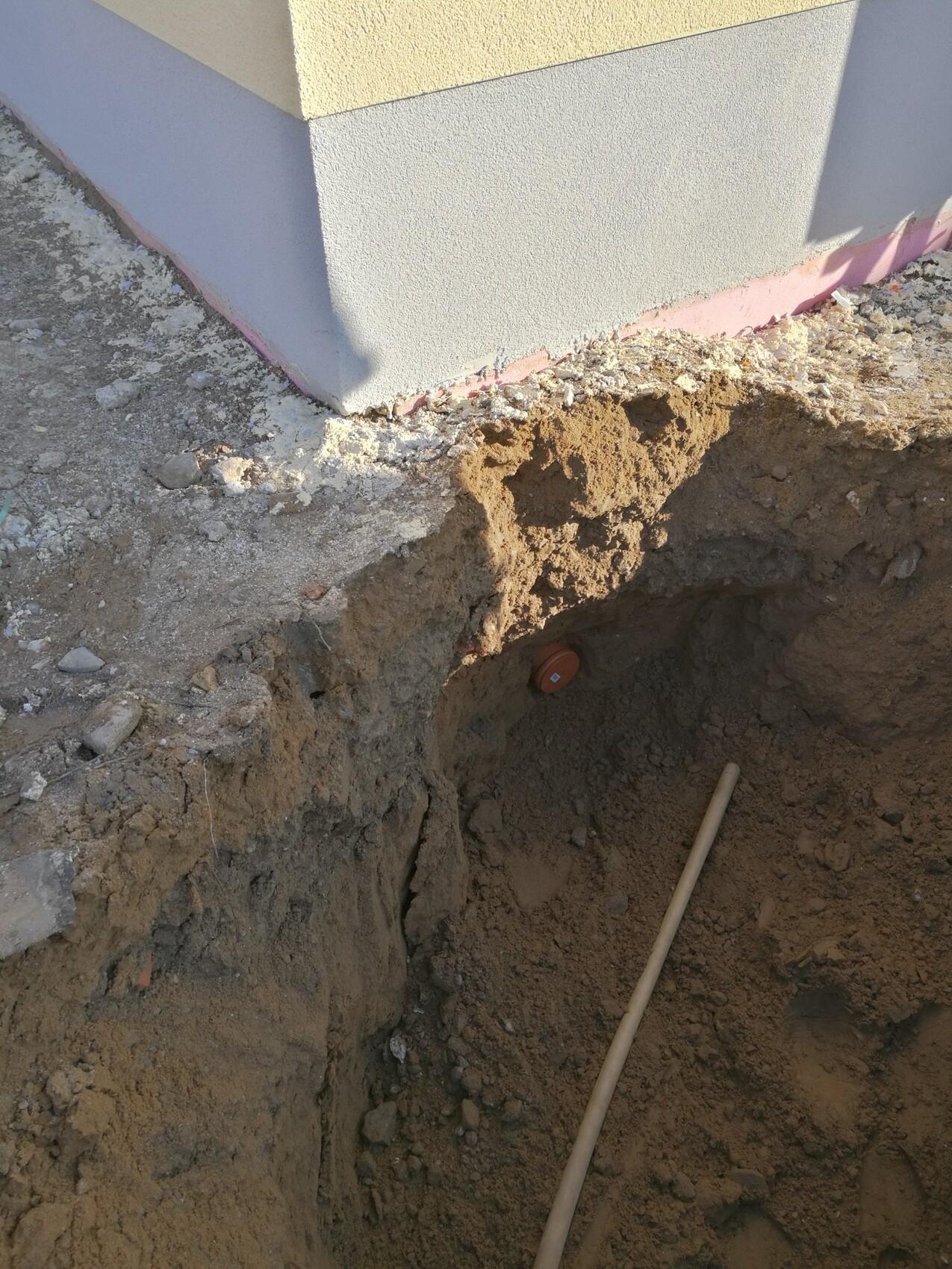 Offene Baugrube neben Hausfundament mit sichtbarem Rohr und Erdarbeiten.