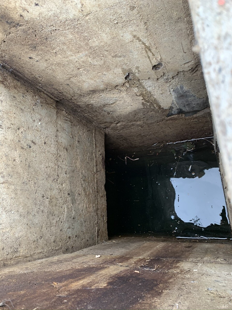 Innenansicht eines rechteckigen Beton-Schachts, rissig und mit stehendem Wasser am Boden.