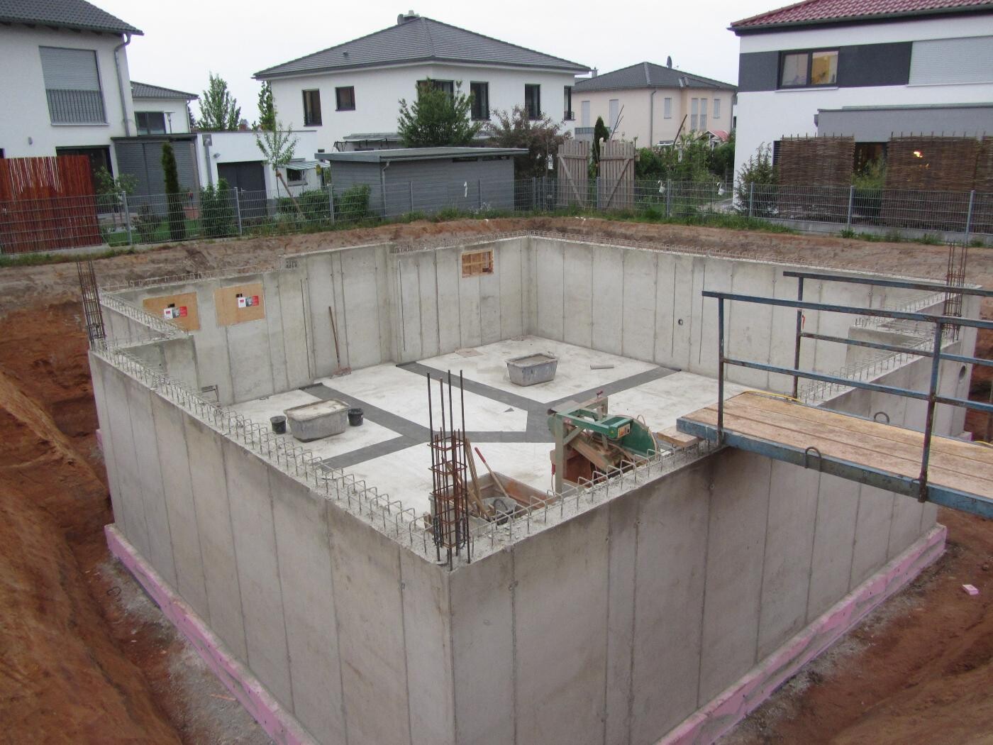 Fundamentgrube aus Beton mit Stahlbewehrung; Wohnhäuser im Hintergrund.