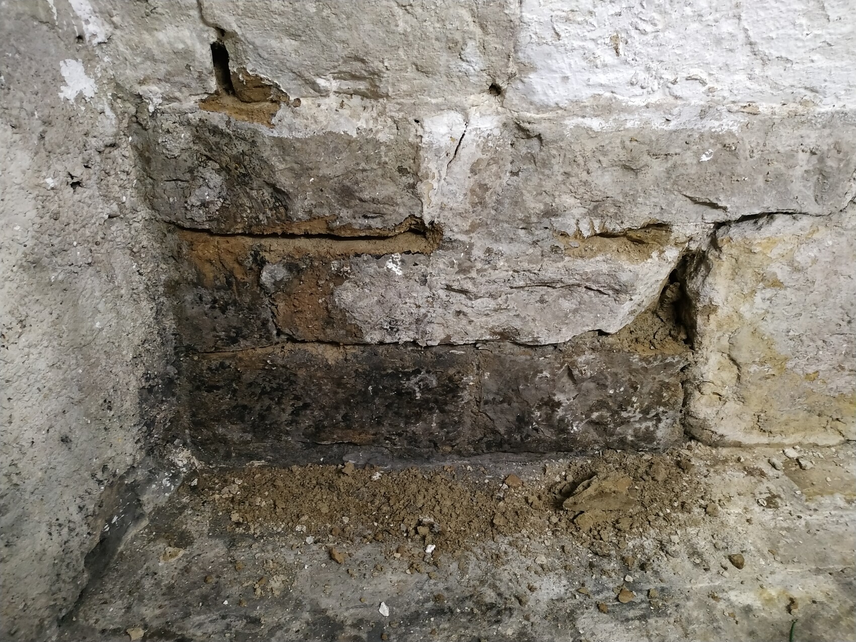 Beschädigte Stein- und Ziegelwand mit dunklem unteren Bereich und Erdreich am Boden.