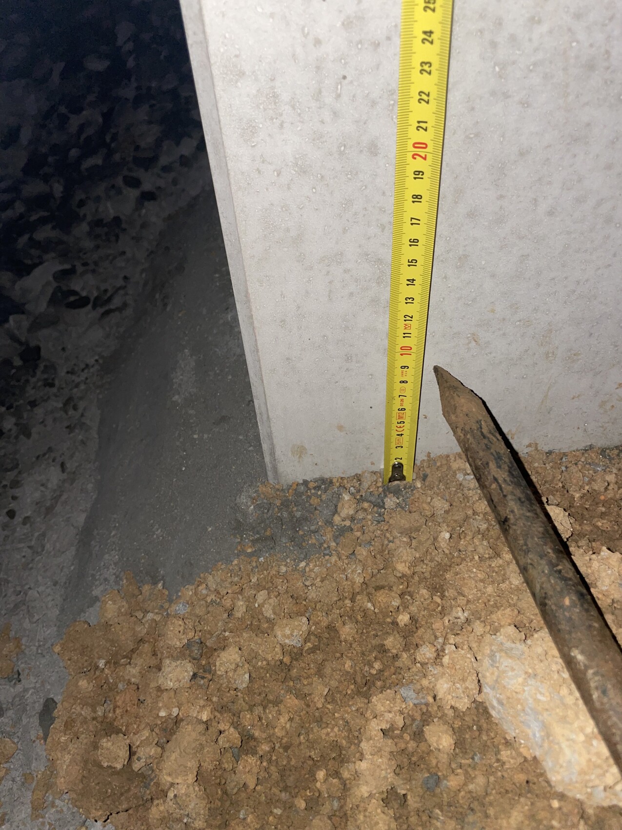 Maßband misst Höhe am rohen Betonfundament, umliegendes Erdreich sichtbar.