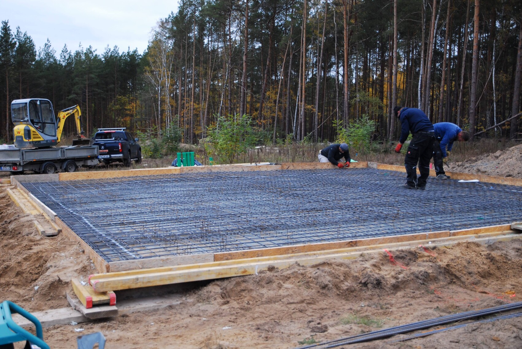 Fundamentbau: Bewehrungsstahlgitter wird verlegt; drei Arbeiter arbeiten, Wald im Hintergrund.