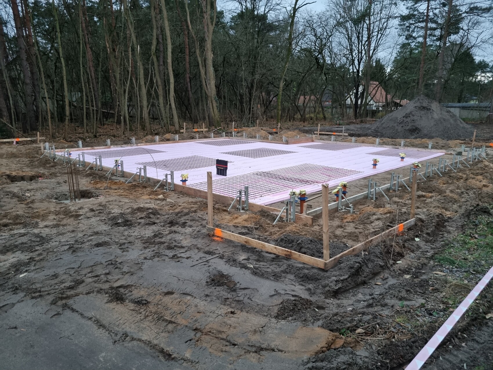Fundamentplatte auf rosa Dämmplatten mit Bewehrungsgitter, Holzrand und Erdarbeiten.