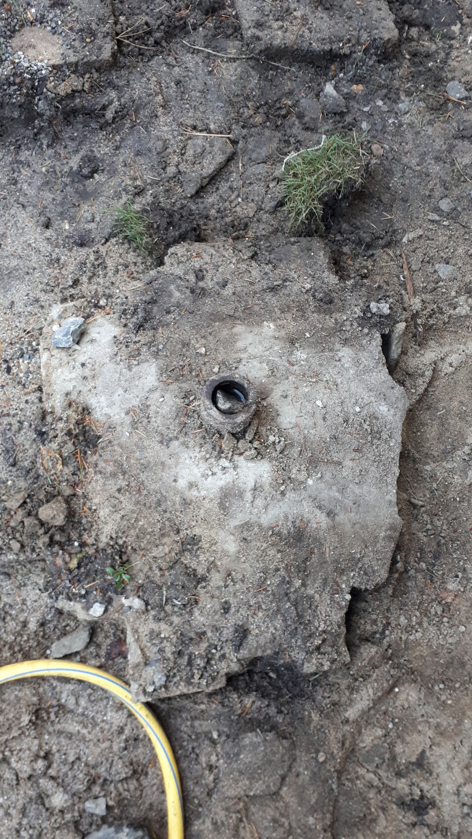 Betonplatte mit runder Öffnung, vermutlich Rohr, umgeben von Erde, Kies und Gras.