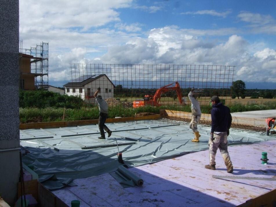 Bauarbeiter verlegen Folie auf einer Baustelle; Bagger im Hintergrund.