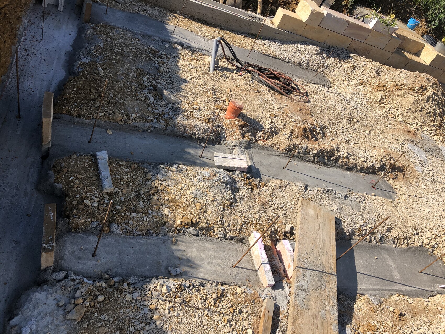 Baustelle mit Kies, Betonfundamenten, Bewehrungsstangen und Holzbalken.