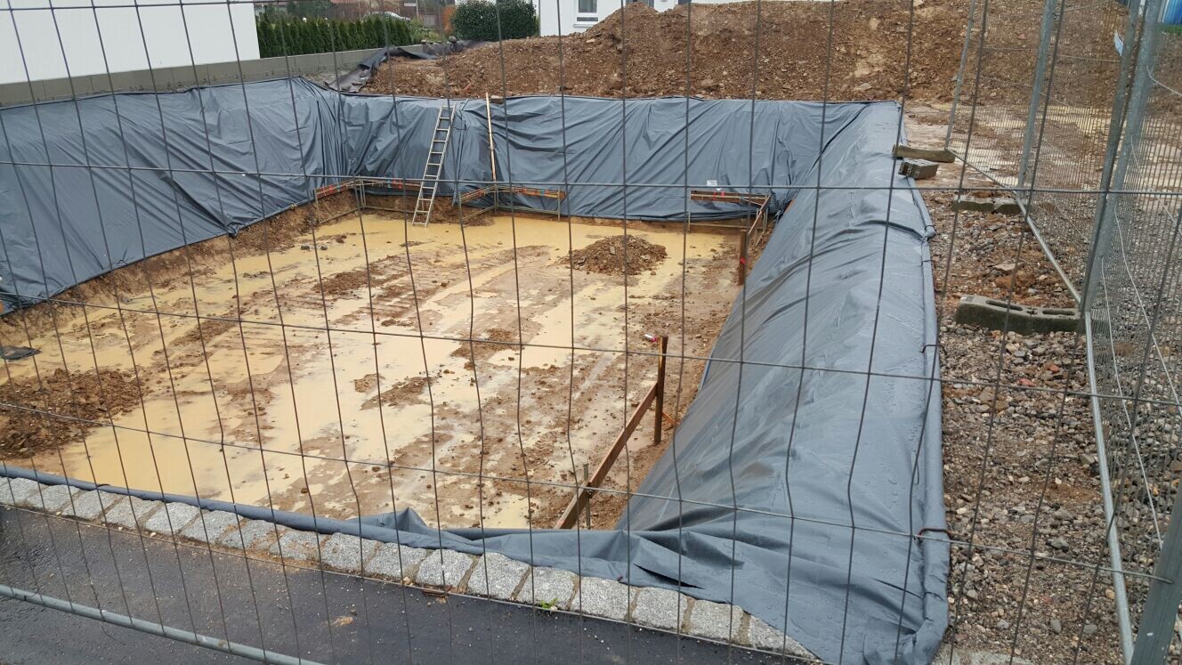 Baustelle: Baugrube mit grauer Plane umrandet; Leiter an Erdwand, matschiger Boden.