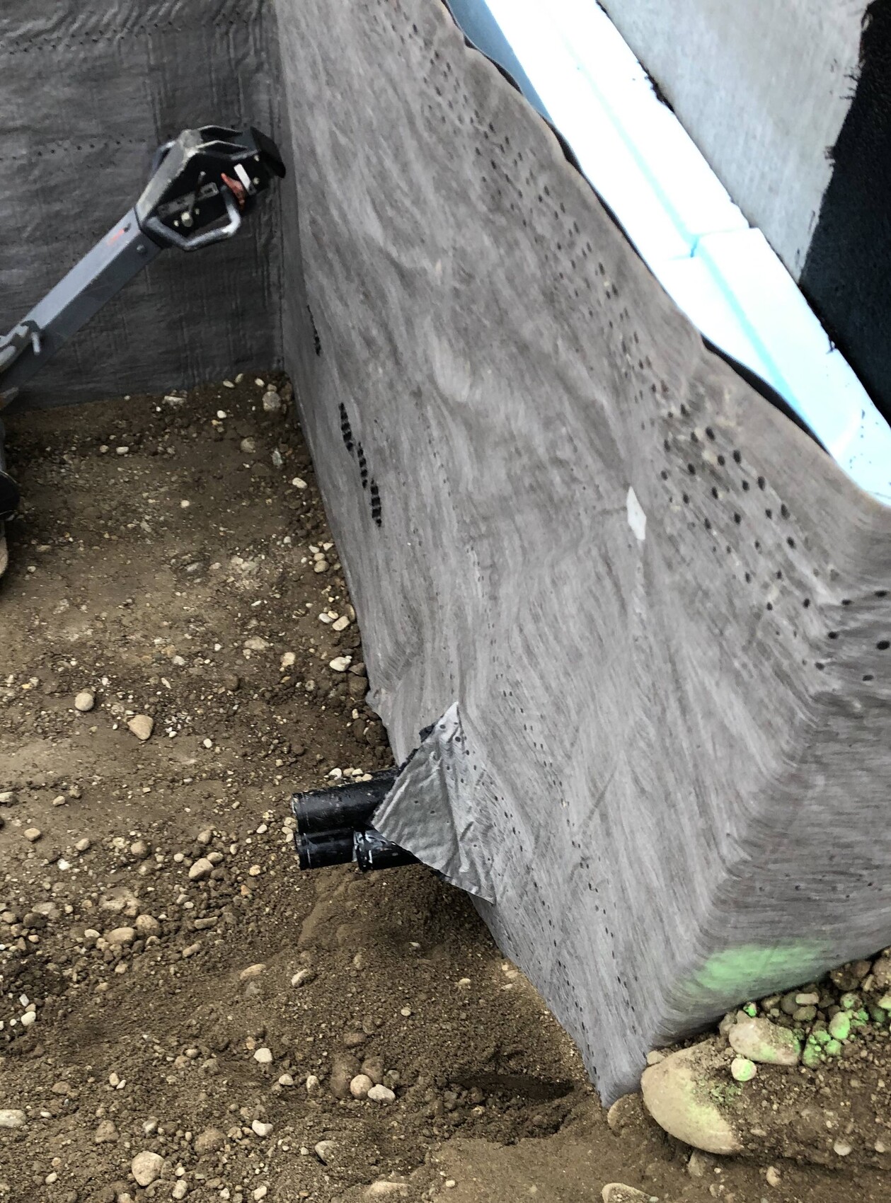 Graue Bau-Linerwand in einer Baugrube; aus dem Boden ragt ein schwarzes Rohr, Kies und Erde.