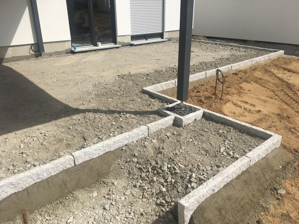 Gartenbaustelle: Steinbegrenzte Beete, zentraler Metallpfosten im Erdreich, Hausfront im Hintergrund.