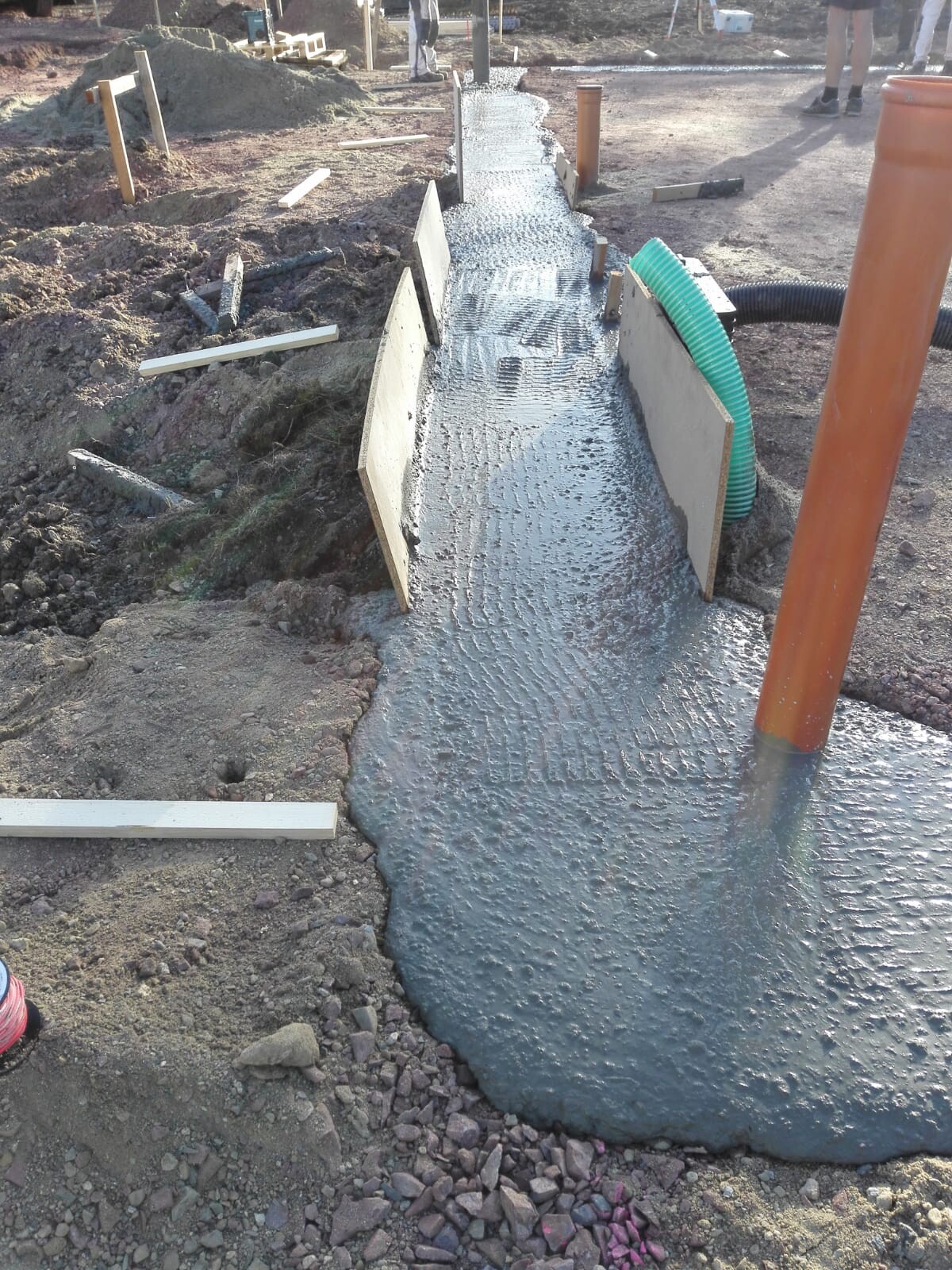 Frisch gegossene Fundamentplatte auf Baustelle mit grünem Schutzschlauch, orangem Rohr und Holzbohlen
