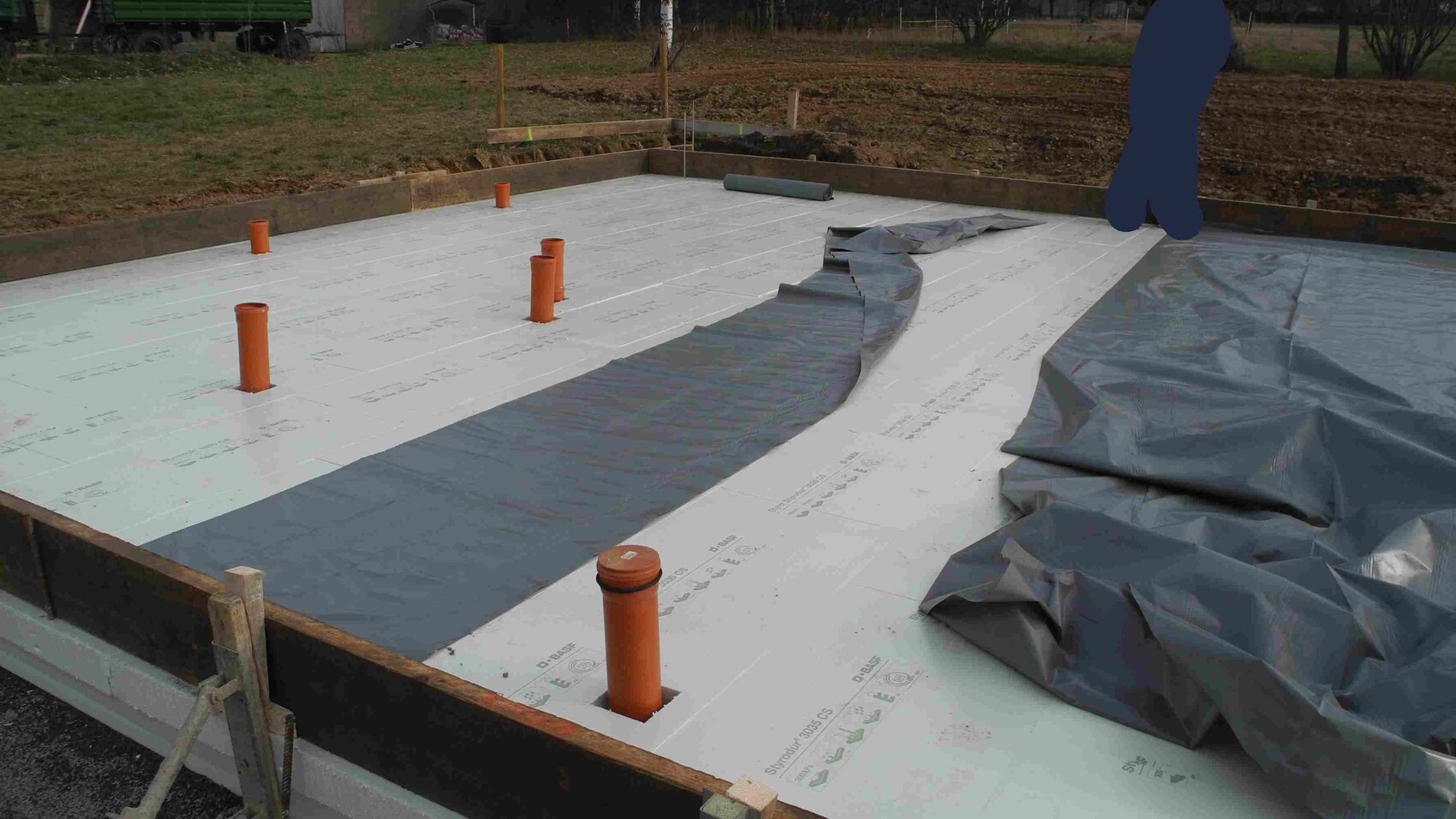 Baustellenfundament mit weißen Dämmplatten, orange Rohre ragen; graue Folie.